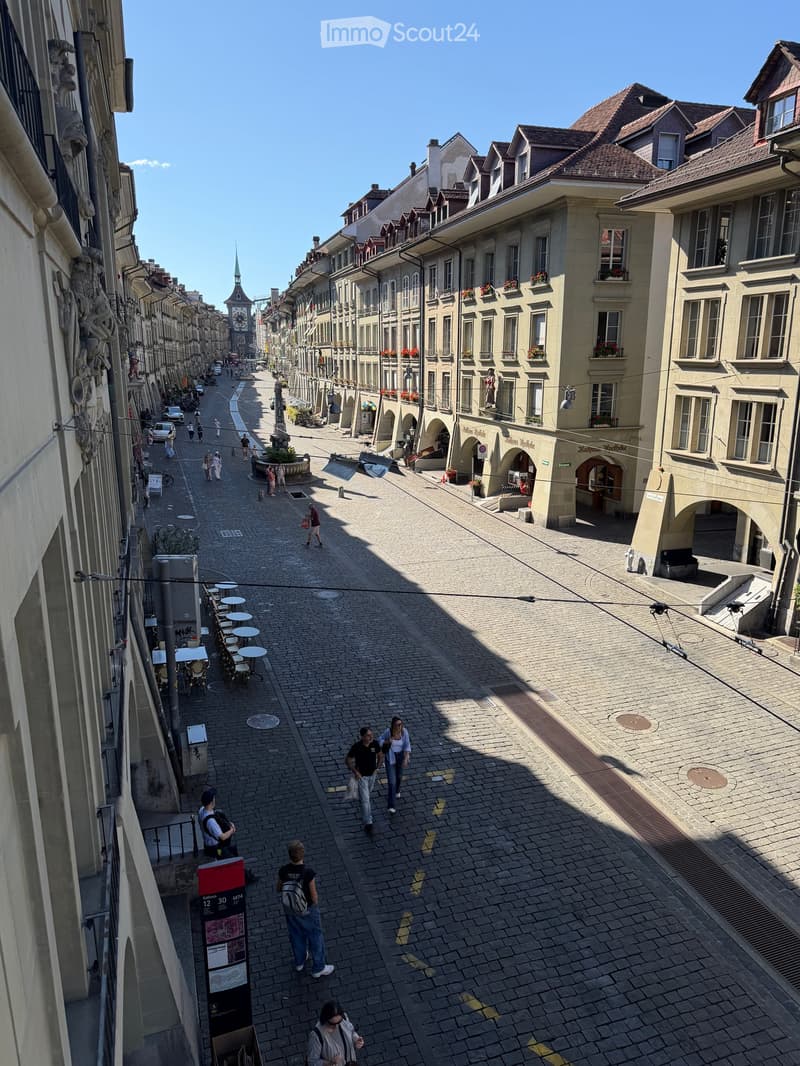 A wide, cobblestone street lined with historic buildings, featuring balconies with flower boxes, pedestrians, and outdoor seating areas.