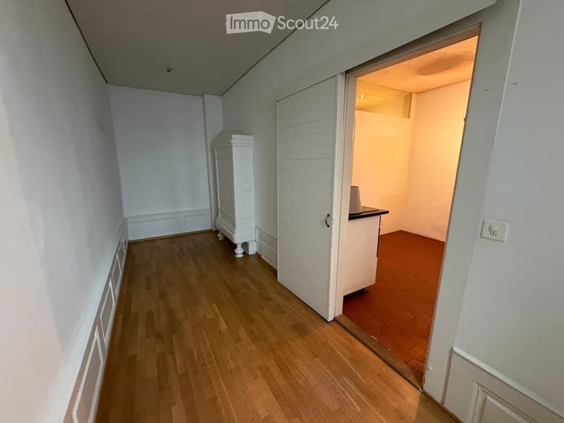 A narrow hallway with wooden flooring, white walls, and a partially open door leading to another room with a visible counter and lamp.