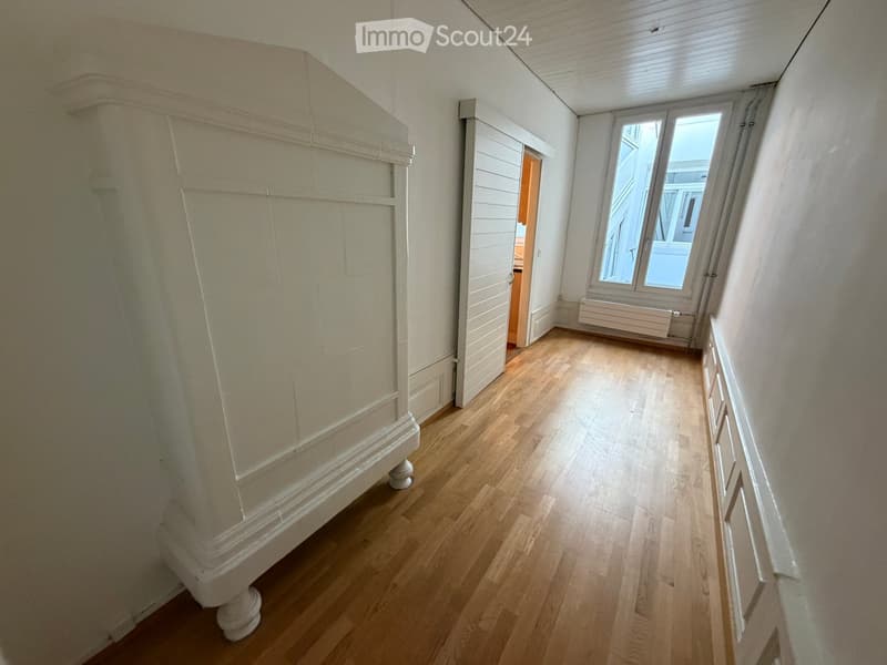 A narrow hallway with wooden flooring, white walls, and a large window at the end. A white radiator is visible beneath the window, and a door leads to another room.