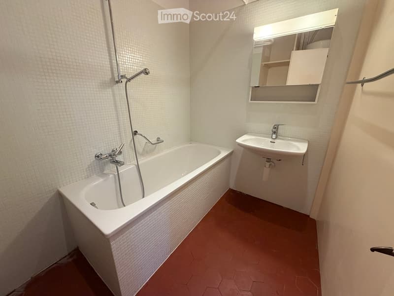 Modern bathroom with a white bathtub, a sink, and a mirror with built-in storage. The walls are tiled, and the floor is covered with hexagonal tiles.