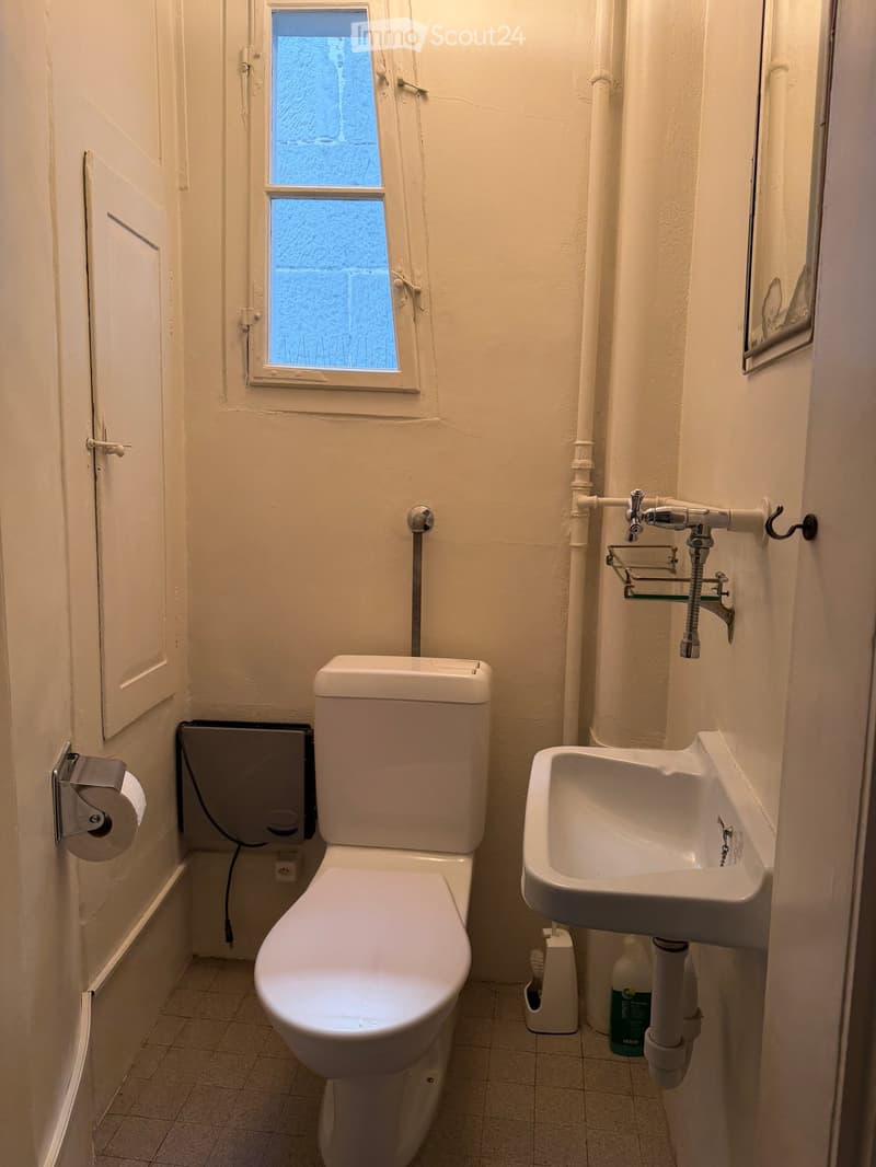 Small bathroom with a toilet, sink, and window. Includes a toilet paper holder, toothbrush holder, and a mirror above the sink.