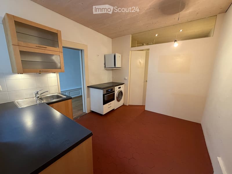 Modern kitchen with dark countertops, wooden cabinets, stainless steel sink, oven, and washing machine. The space features a red hexagonal tile floor and white walls.