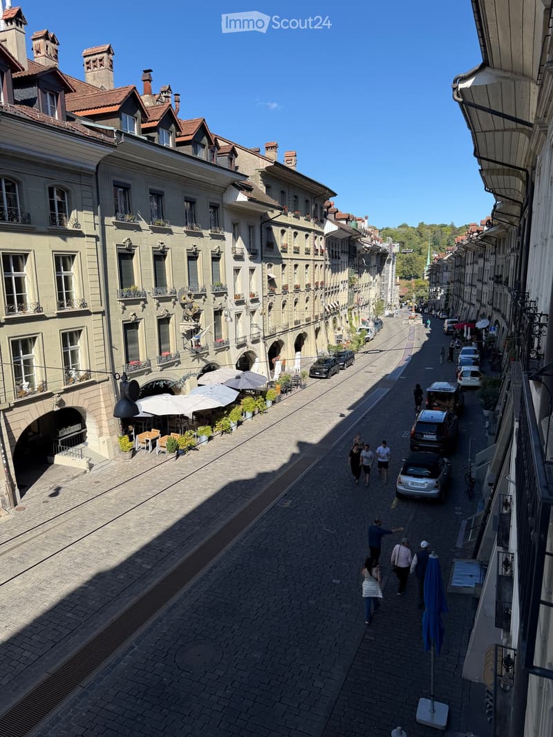 A picturesque European street lined with multi-story buildings featuring balconies, large windows, and ornate facades. The street is paved with cobblestones and has outdoor seating areas with umbrellas and tables, with pedestrians walking along the sidewa