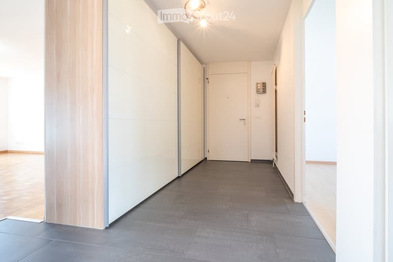 Modern hallway with grey tiled floor, white walls, and wooden paneling on one side. Features a closed door and recessed lighting fixtures.
