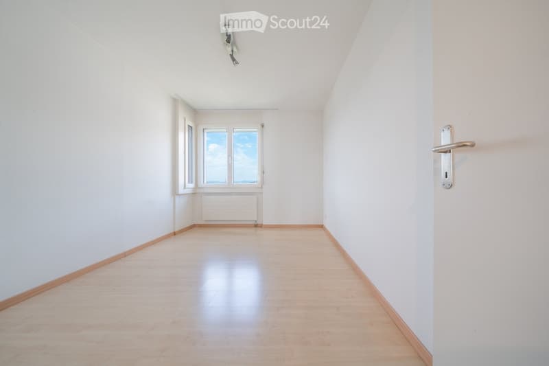 Empty room with light wooden flooring, white walls, and large windows letting in natural light. The room has a simple, clean design with a door on the right side.