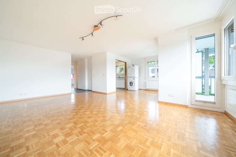 Empty room with wooden herringbone flooring, white walls, large windows, and a washing machine in the background.