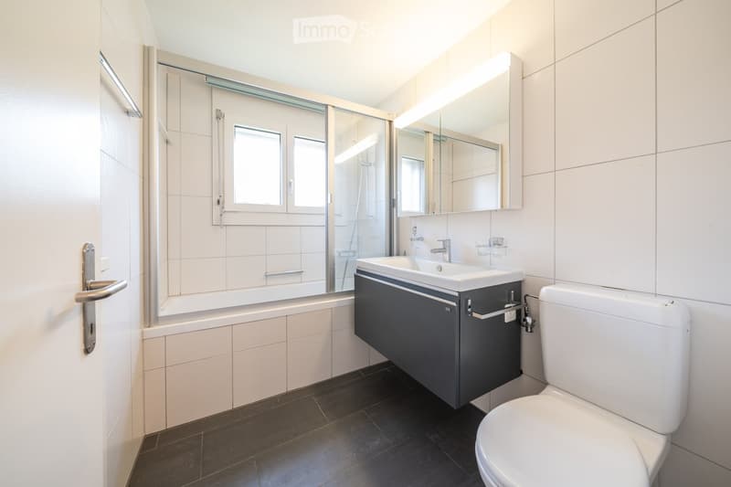 Modern bathroom with a bathtub, sink, toilet, and a large mirror above the sink. The room features white tiled walls and a dark tiled floor.