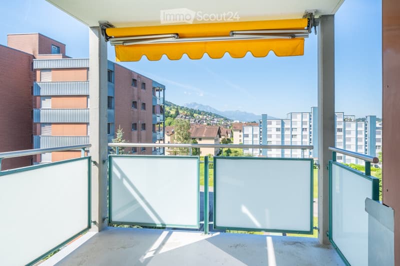 Balcony with metal railings, glass panels, and an orange awning, overlooking a town with buildings and mountains in the background.