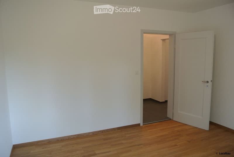 Empty room with wooden flooring and white walls, featuring an open doorway leading to another room.