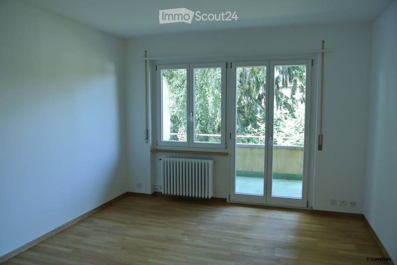 Empty room with wooden flooring, white walls, large sliding glass doors leading to a balcony, radiator, and visible electrical outlets.