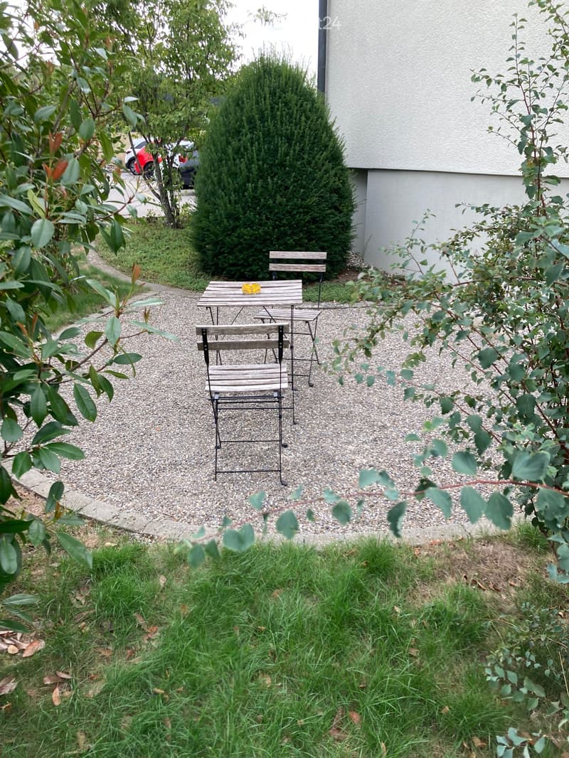Small outdoor seating area with a wooden table and chairs, surrounded by gravel and greenery, with a building wall in the background.
