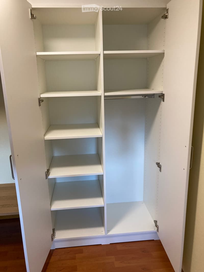 Empty white built-in closet with multiple shelves and a hanging rod, set in a room with wooden flooring.