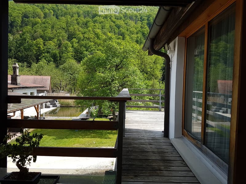 Wooden balcony with a railing, overlooking a lush green landscape and a body of water.