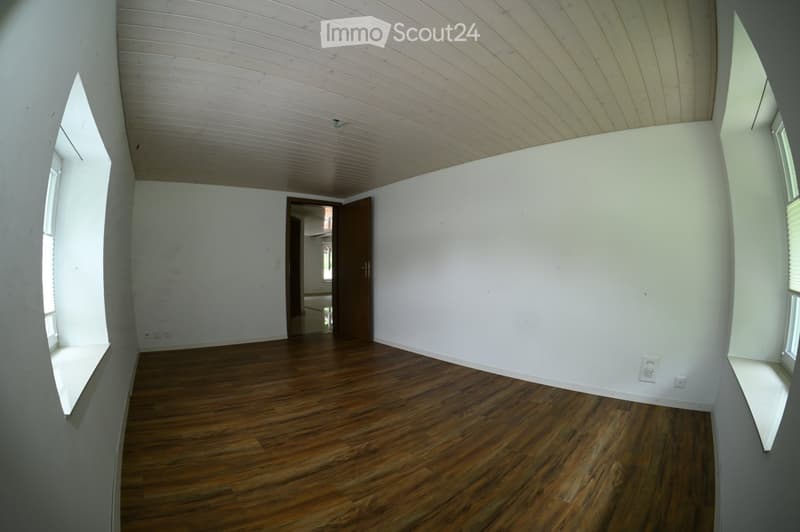 Empty room with wooden flooring, white walls, and a wooden door leading to another space. The ceiling has a paneled design, and there are windows on two walls.
