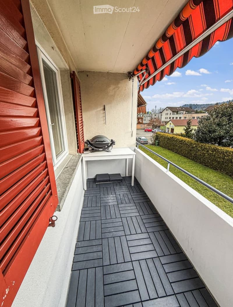 Balcony with grey tiled floor, white table, grill, red shutters, and a view of the neighborhood.