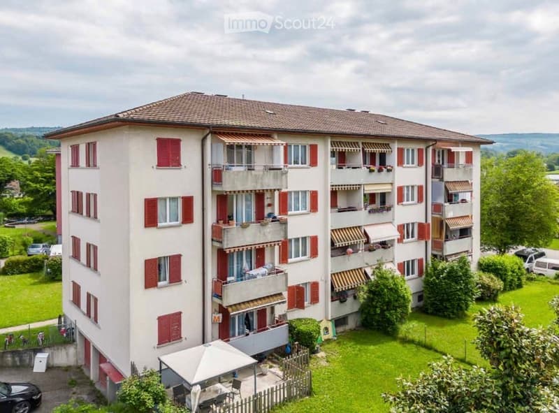 Multi-story residential building with red shutters, balconies, and surrounding greenery.