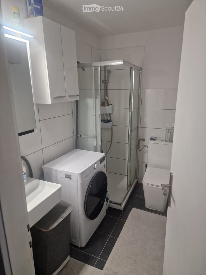 Modern bathroom with a white sink, toilet, glass-enclosed shower, and a washing machine. The space features white cabinets and a dark tiled floor.