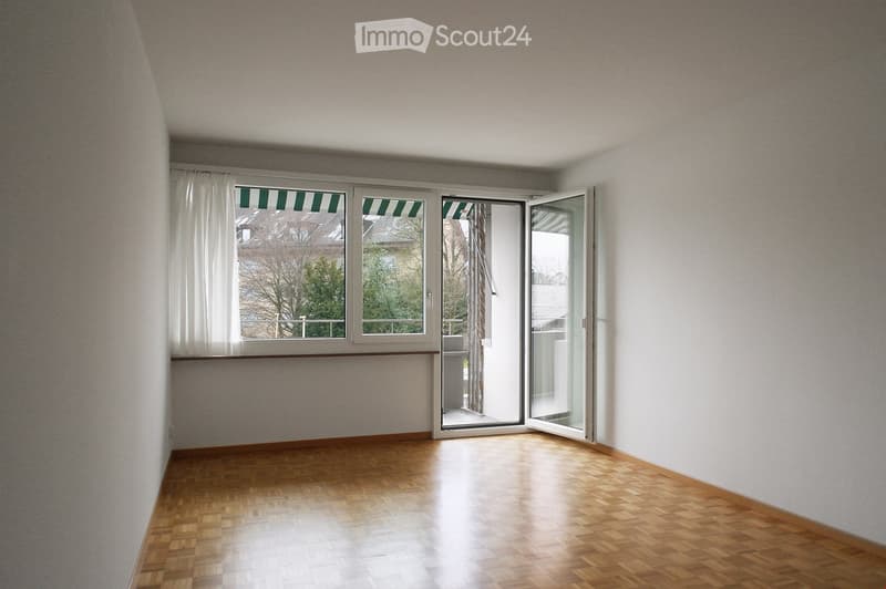 Empty room with wooden flooring, white walls, large windows with green blinds, and an open sliding door leading to a balcony.