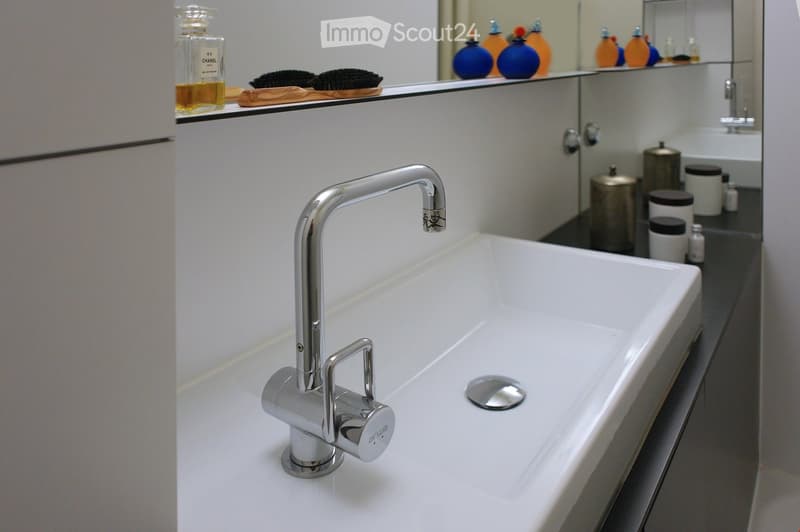 Modern bathroom sink with a sleek chrome faucet, a large white sink basin, and a mirror above it. The mirror has decorative bottles and brushes on the shelf, and the sink has a modern design with a built-in drain.