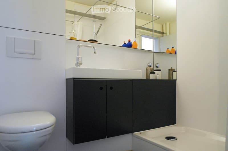 Modern bathroom with white walls, a dark grey cabinet, a white sink, a mirror, a toilet, and a bathtub. The sink has a modern faucet, and the mirror has shelves with decorative bottles and a brush.