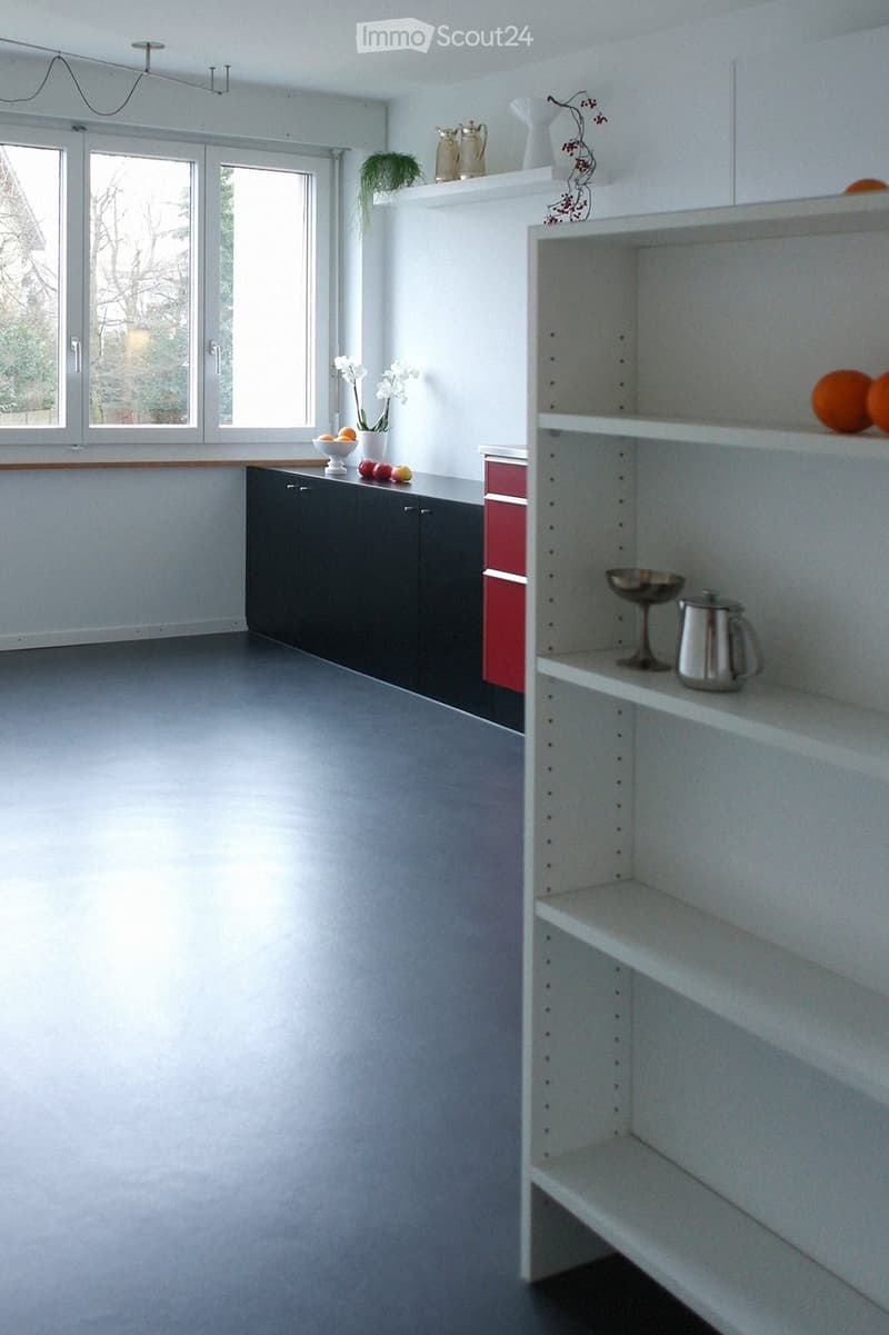 Empty room with white walls, grey floor, large windows, white shelving unit, and a dark cabinet.