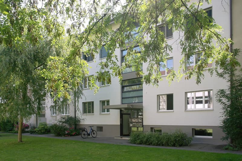 Modern multi-story residential building with white facade, large windows, and greenery surrounding the entrance area.