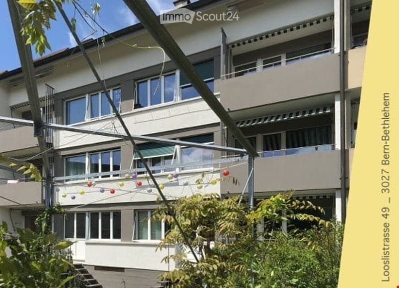 Multi-story residential building with large windows, balconies, and greenery in the foreground.