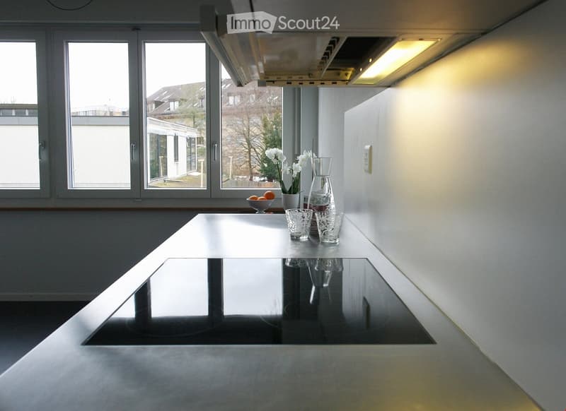 Modern, minimalist kitchen with a sleek, reflective table, glassware, a vase with white flowers, and a bowl of fruit on the counter. Large windows provide natural light and a view of the outdoor area.