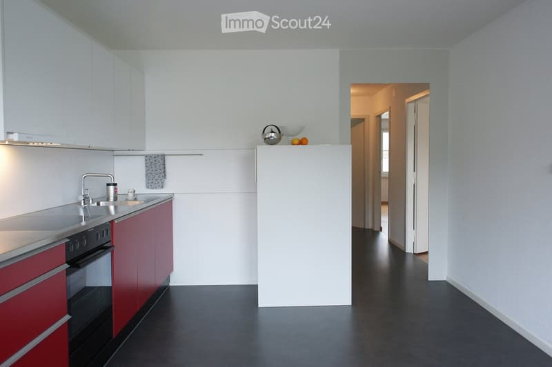 Modern kitchen with red and white cabinets, stainless steel sink, oven, and a small refrigerator. The floor is dark, and the walls are white. A teapot and a cup are placed on the counter.