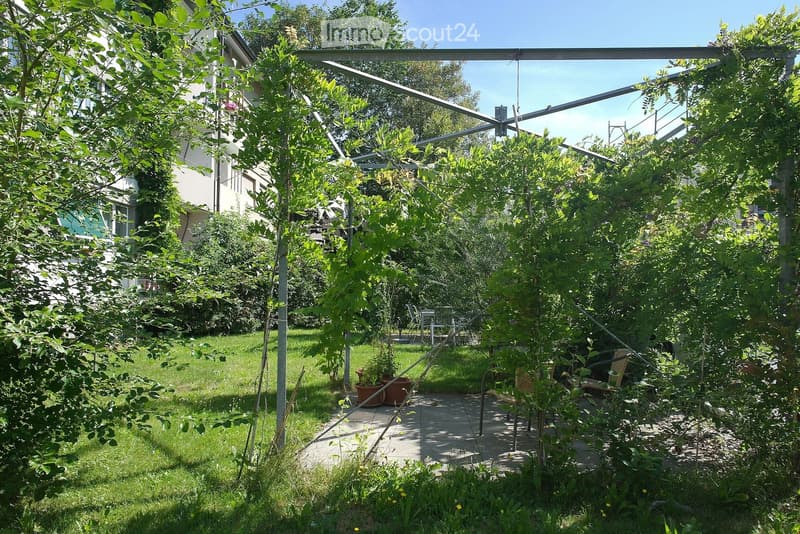 A lush garden with a pergola structure, surrounded by trees and greenery, with a patio area and outdoor furniture visible in the background.