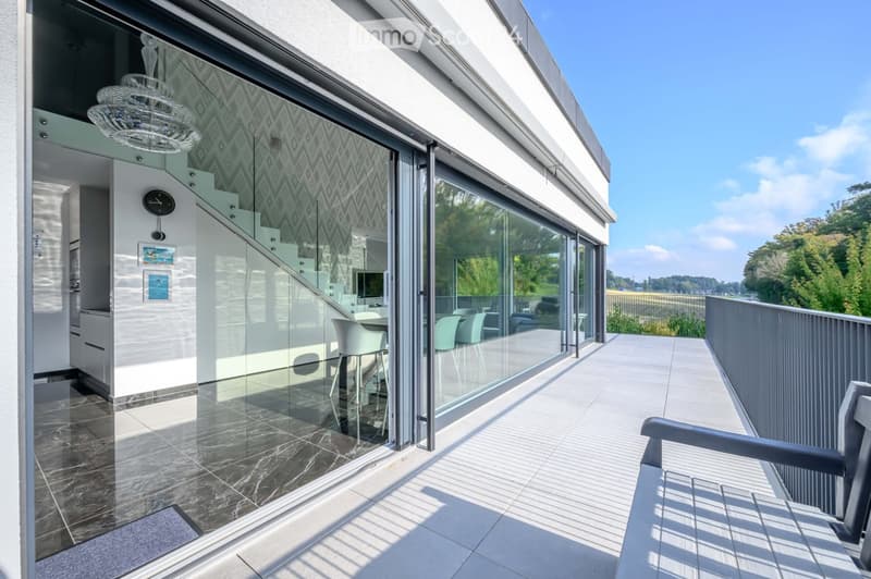 Balcony / Terrace. Modern balcony with glass walls, tiled floor, and metal railings. Features a chandelier, a table with chairs, and a bench. The interior reflects through the glass, showing a staircase and kitchen.