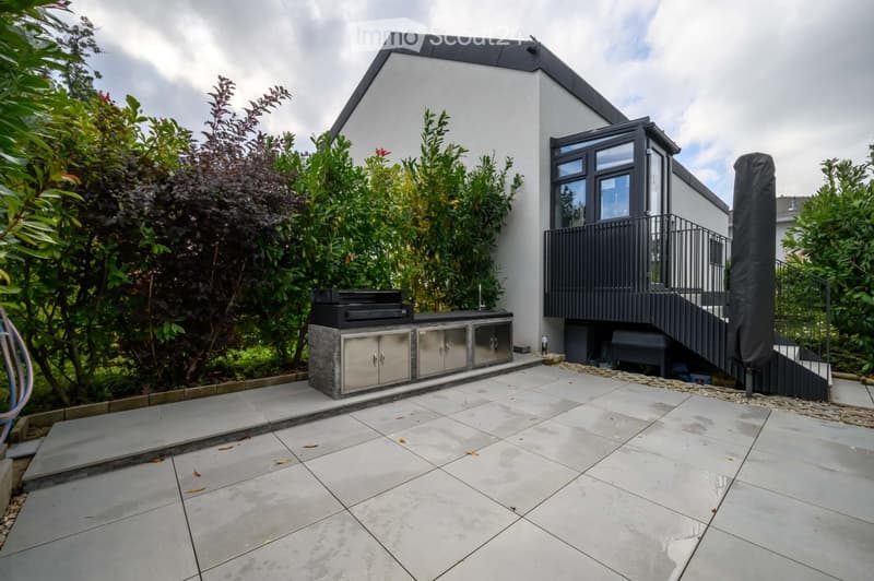 Garden / Outdoor Area. Outdoor area with a modern outdoor kitchen, grill, and tiled floor. Stairs lead to a covered area with metal railings. The area is surrounded by plants and a fence.