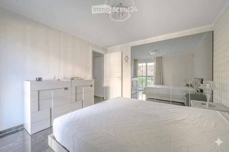 Bedroom. Spacious bedroom with a large bed, white walls, and a mirrored wardrobe. The room features a modern chandelier and a nightstand with a lamp.