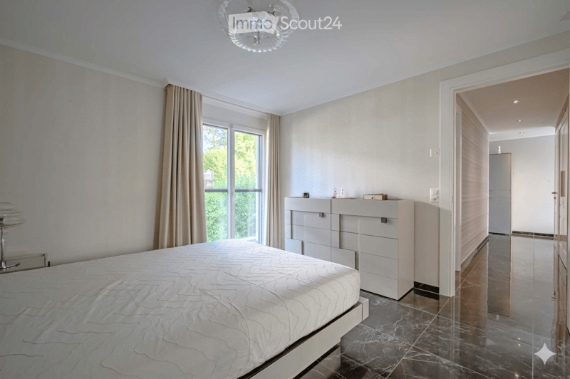 Bedroom. Spacious bedroom with a large bed, white walls, and marble flooring. A nightstand with a lamp is on the left, and a set of drawers is against the wall. The room has a large window with curtains.