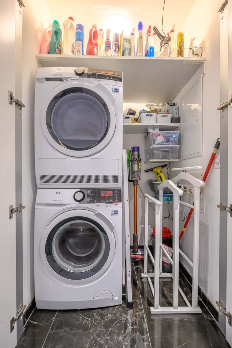 Furnished Room. A small laundry room with a stacked washer and dryer, a vacuum cleaner, and various cleaning products on the shelves.