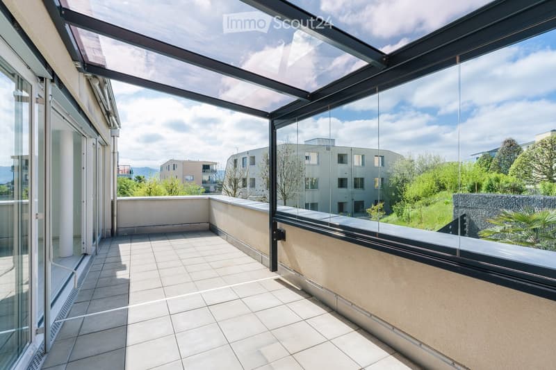 Modern balcony with glass roof and walls, tiled floor, and a view of surrounding buildings and greenery.