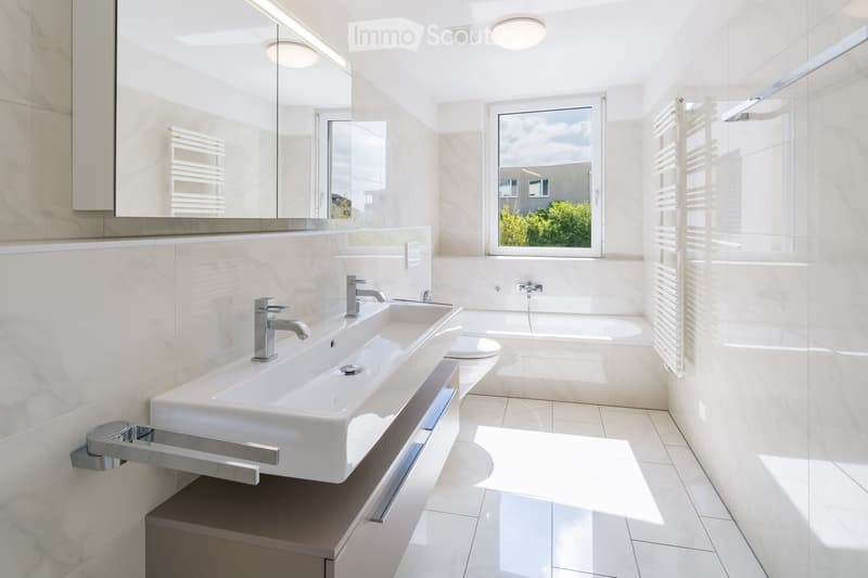 Modern bathroom with a double sink, bathtub, toilet, and large mirror. The room features white marble tiles and a large window letting in natural light.