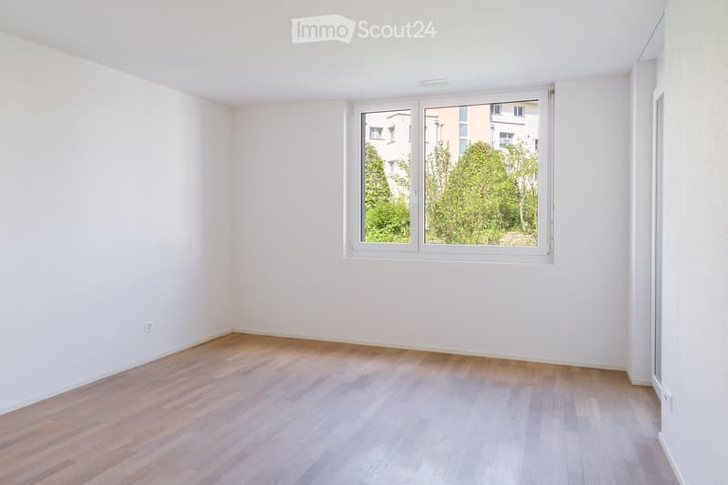 Empty room with light wooden flooring and white walls, featuring a large window with a view of greenery outside.
