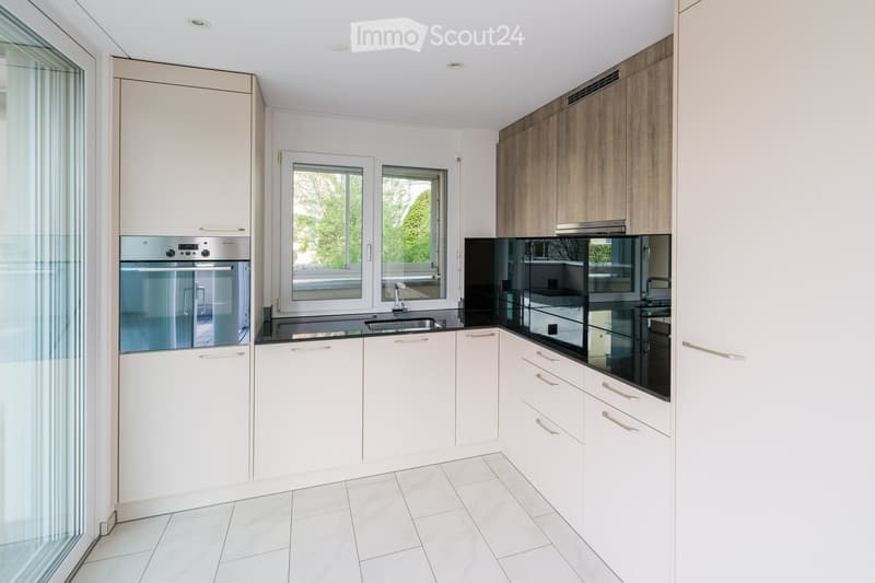 Modern kitchen with white cabinets, stainless steel oven, black countertop, and large windows overlooking a garden.