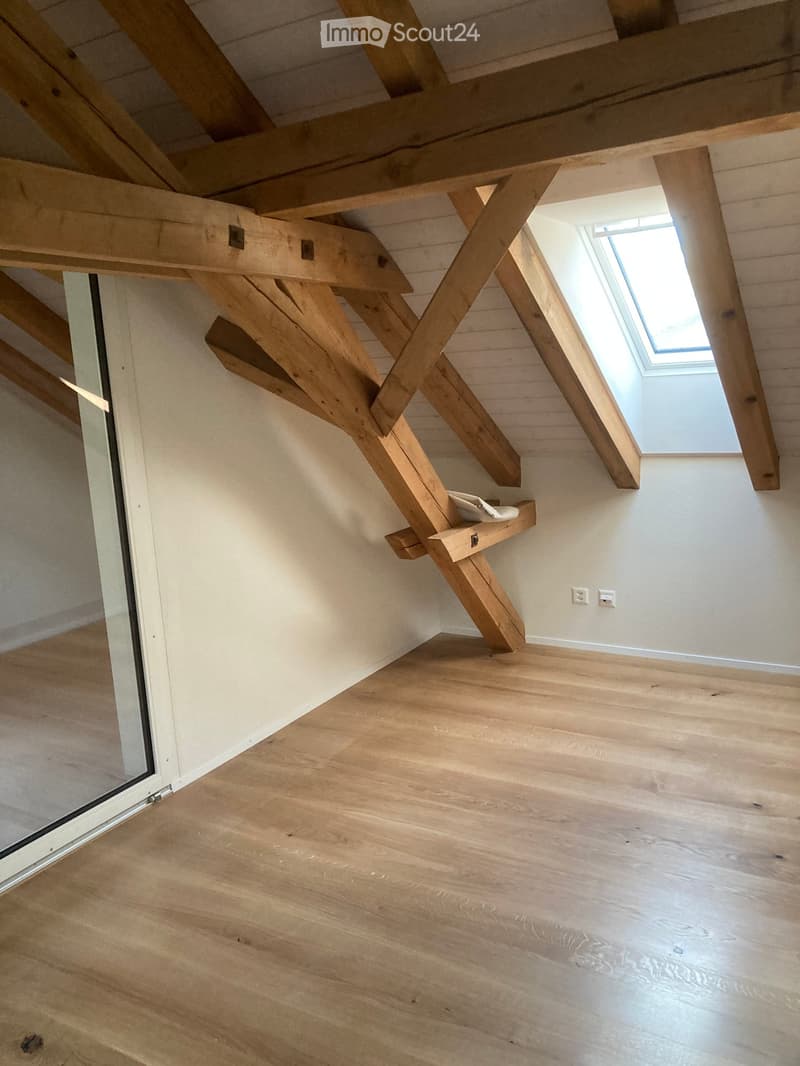 Empty room with wooden floor, white walls, exposed wooden beams, skylight window, and a small wooden shelf with a white bowl on it.