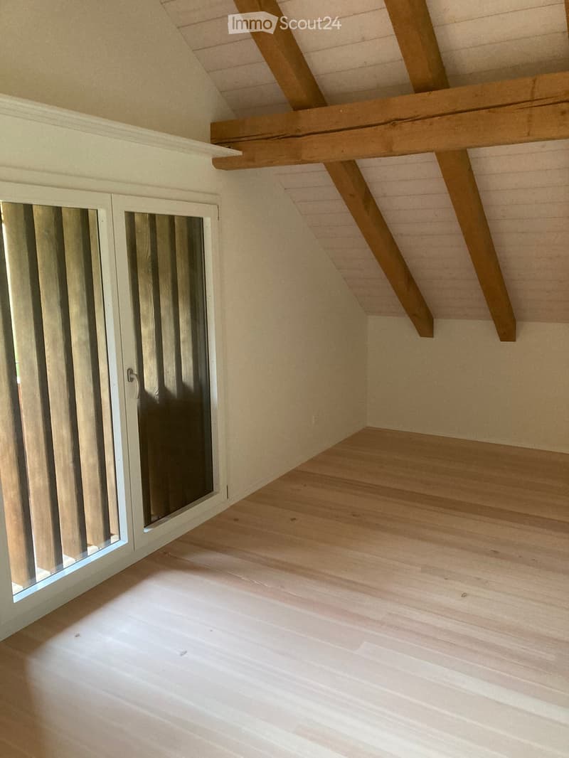 Empty attic space with wooden beams, exposed wooden ceiling, and a small window with white frame and glass panels.