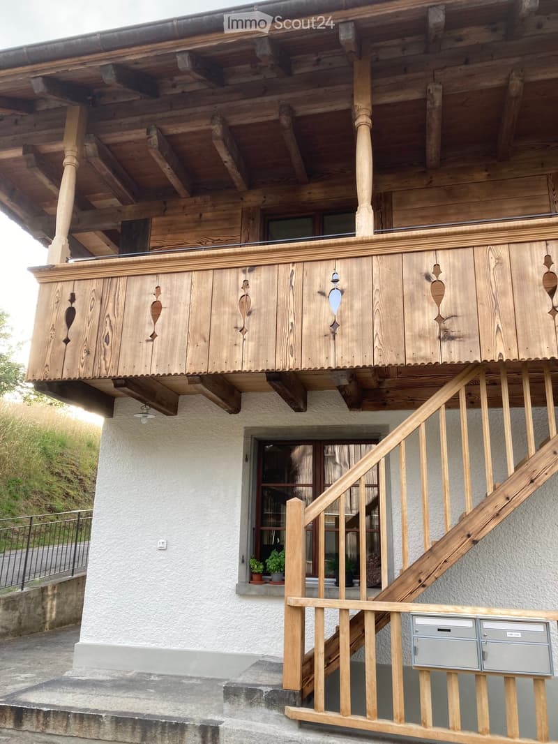 A two-story wooden house with a balcony, wooden staircase, and small potted plants near the entrance.