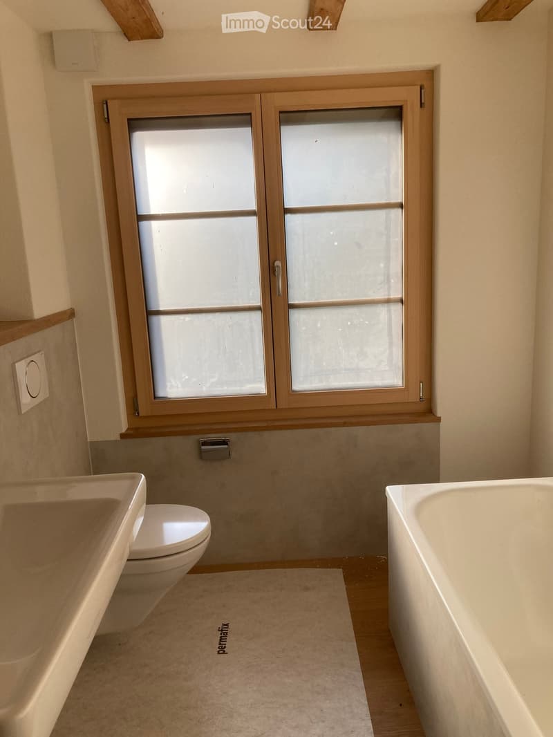 Modern bathroom with a toilet, bathtub, and sink, featuring a large frosted glass window and light-colored tiles.