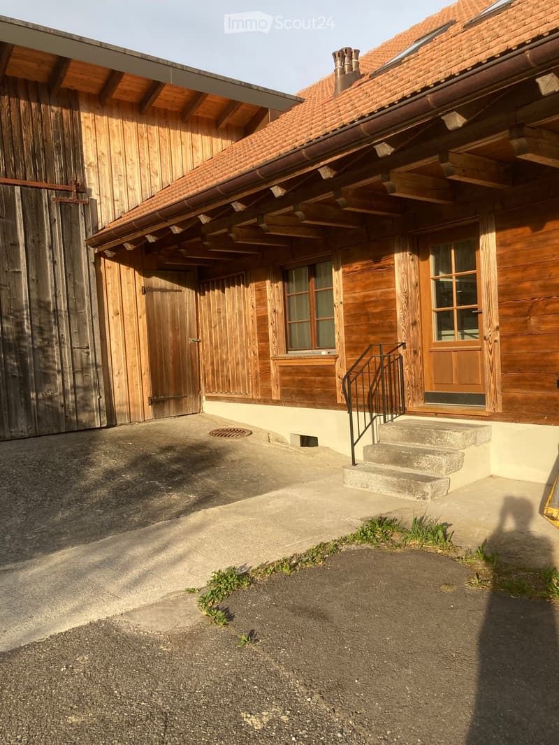A two-story wooden house with a red-tiled roof, multiple windows, and a concrete pathway leading to a staircase with a metal railing.