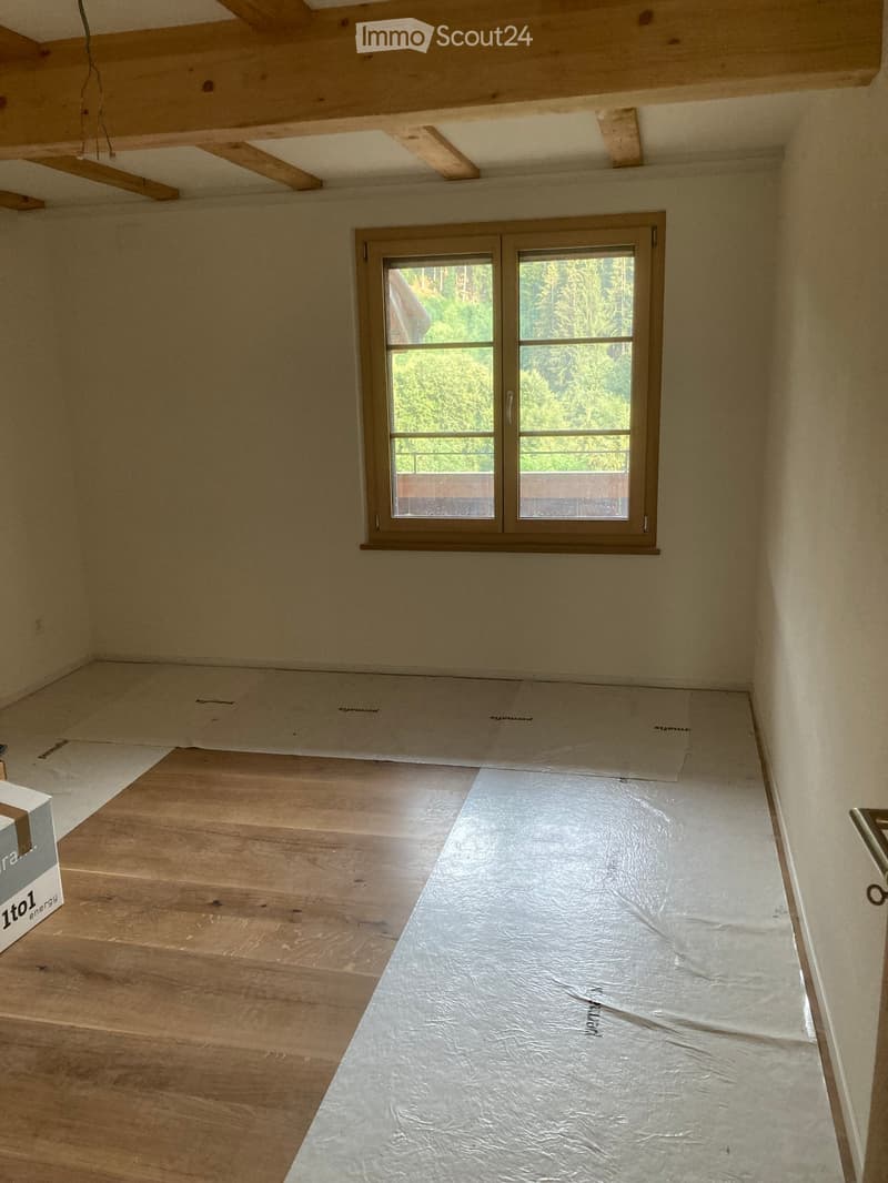 Empty room with wooden flooring, exposed wooden beams on the ceiling, and a large window with a view of greenery.