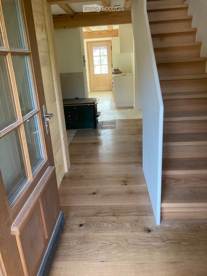 Spacious hallway with wooden flooring, white walls, and a staircase leading upwards. Features a wooden door with glass panels on the left and a glimpse of a room with a green cabinet and a staircase in the background.