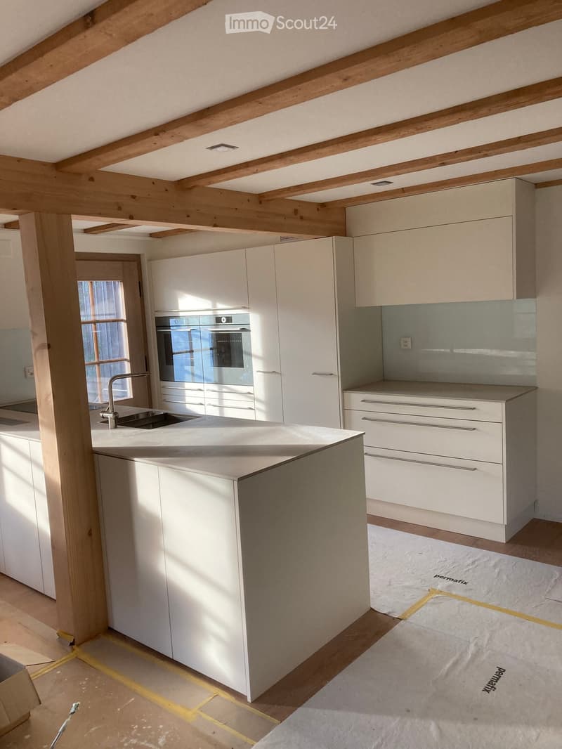 Modern kitchen with white cabinets, a central island, and a stainless steel oven. Exposed wooden beams on the ceiling and a large window provide natural light.