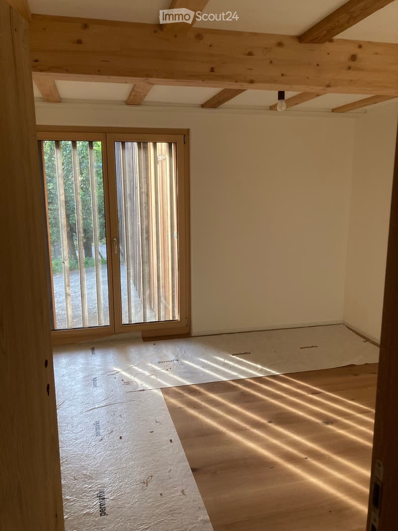 Empty room with wooden floor and exposed wooden beams on the ceiling. Large sliding glass door with wooden frame leading to outdoor area.