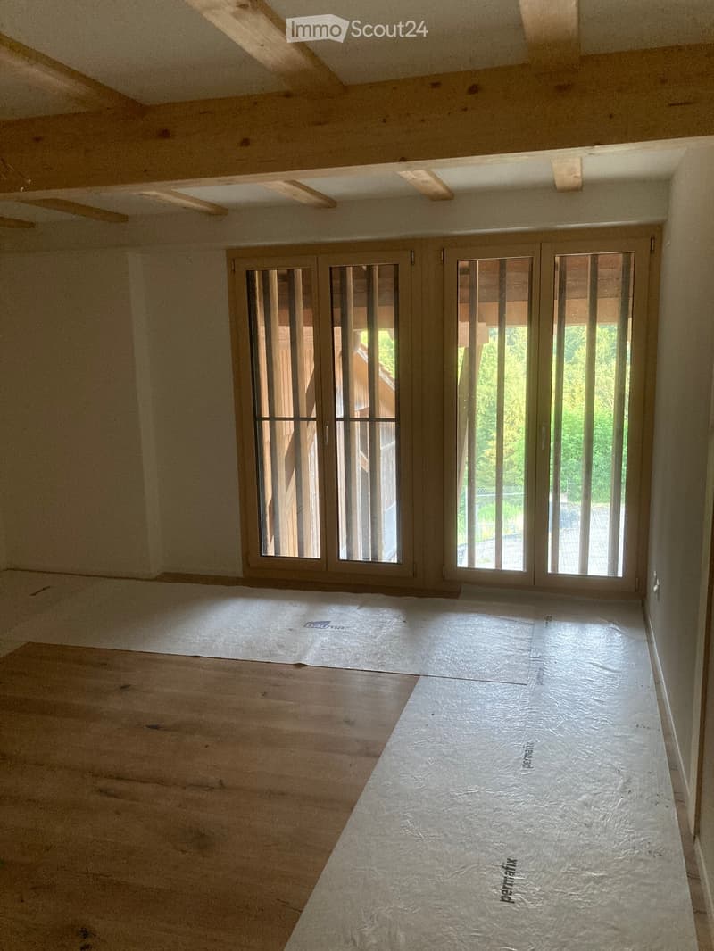 Empty room with exposed wooden beams on the ceiling, unfinished wooden flooring, and large sliding glass doors leading outside.