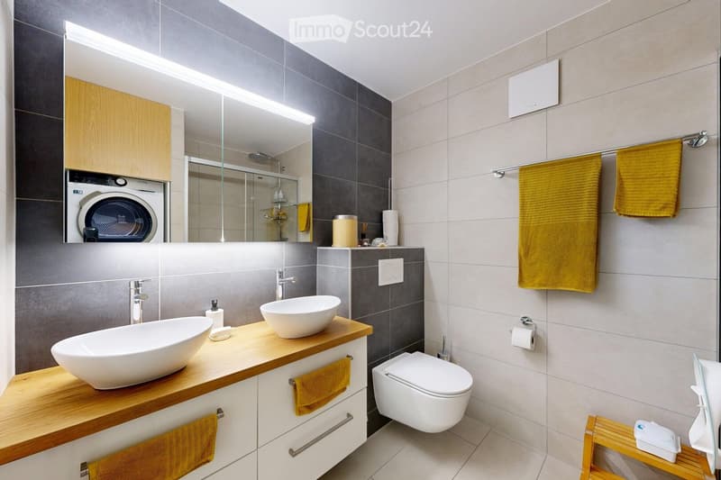 Modern bathroom with a double sink, toilet, large mirror, and built-in washing machine. Features wooden cabinets, grey tiles, and yellow towels.